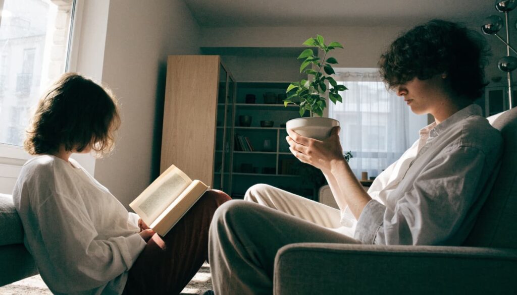 One partner reads while the other tends a houseplant in a bright living room.