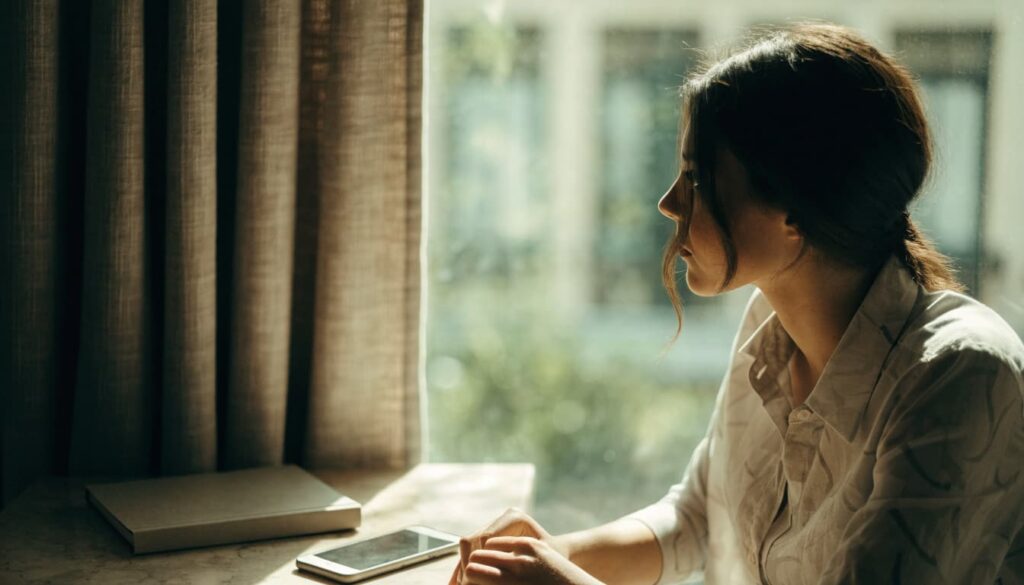 Limerence vs. Love: What the Difference Can and Can’t Tell You 3 Woman sitting beside a window, looking outside with a book and phone on the table.